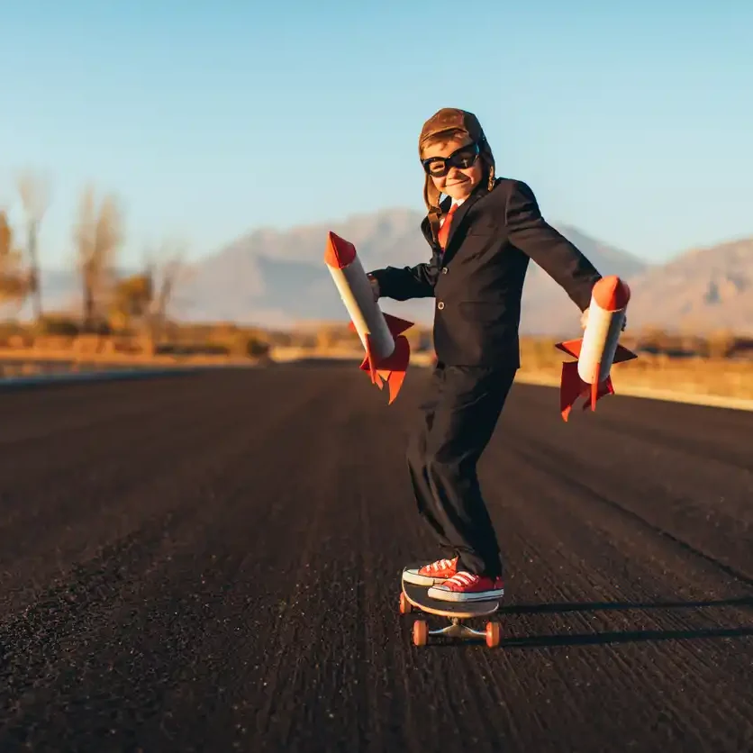 Kid in suit and goggles with small toy rockets in hand rolling down long regional street on skateboard.