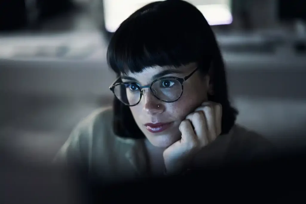 Female SEO AI expert with glasses and hand on chin in front of computer screen, bright lights lighting up her face.