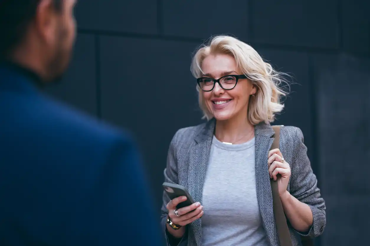 Blonde businesswoman smiling on street at copywriter colleague with phone in her hand