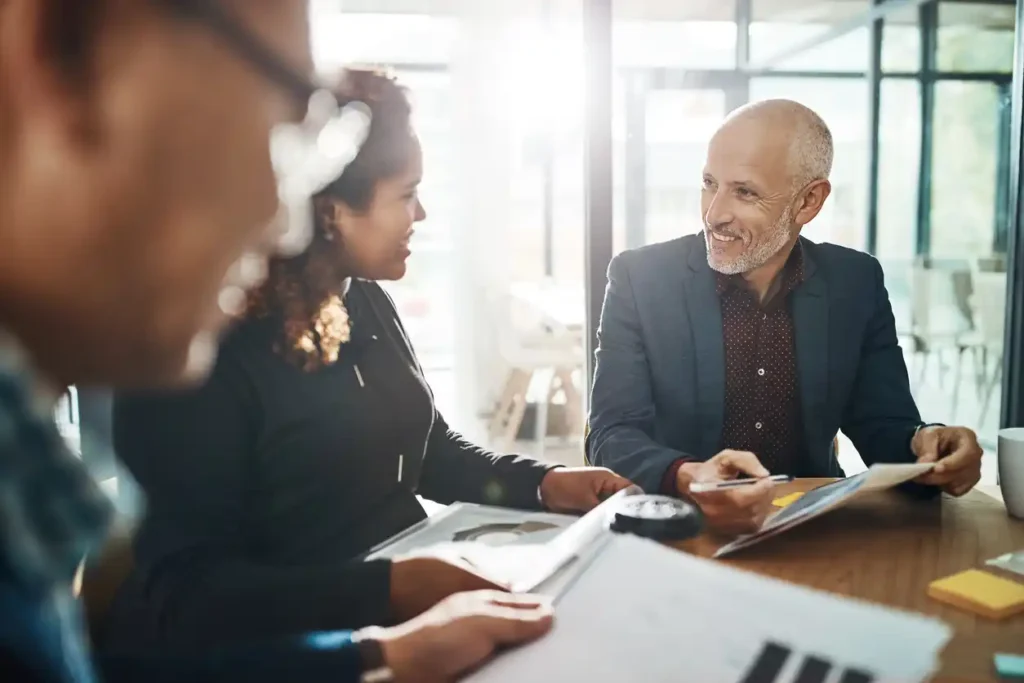 Mortgage broker discussing home loan application with leads at a boardroom table, sunlight flowing from the background window.