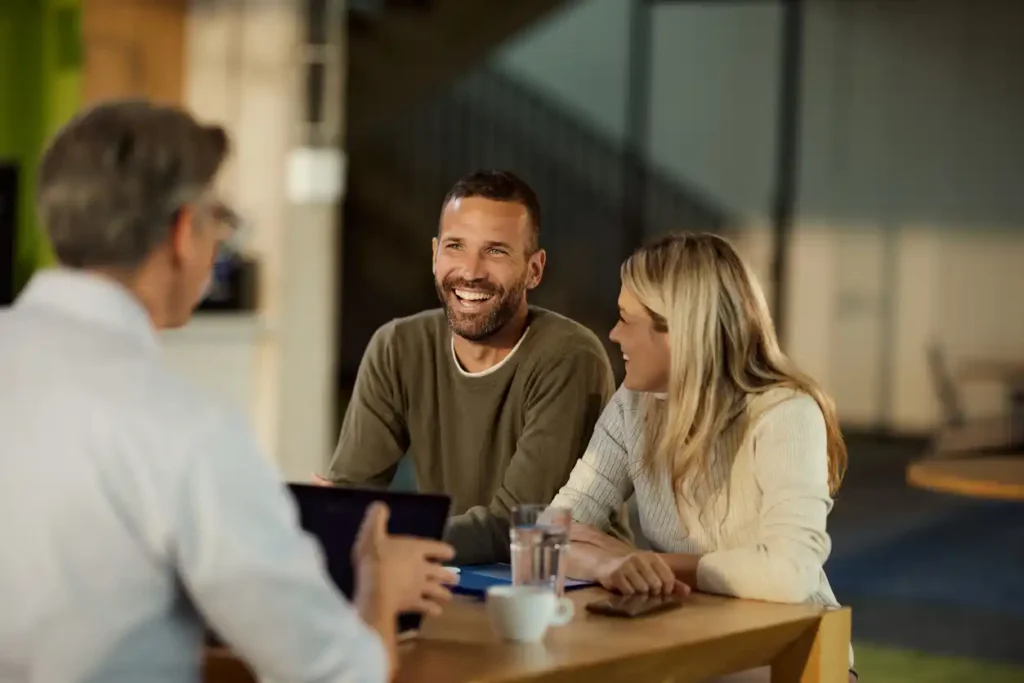 Mortgage broking clients - a young vibrant couple - at a desk alongside their finance expert in a warmly lit room.
