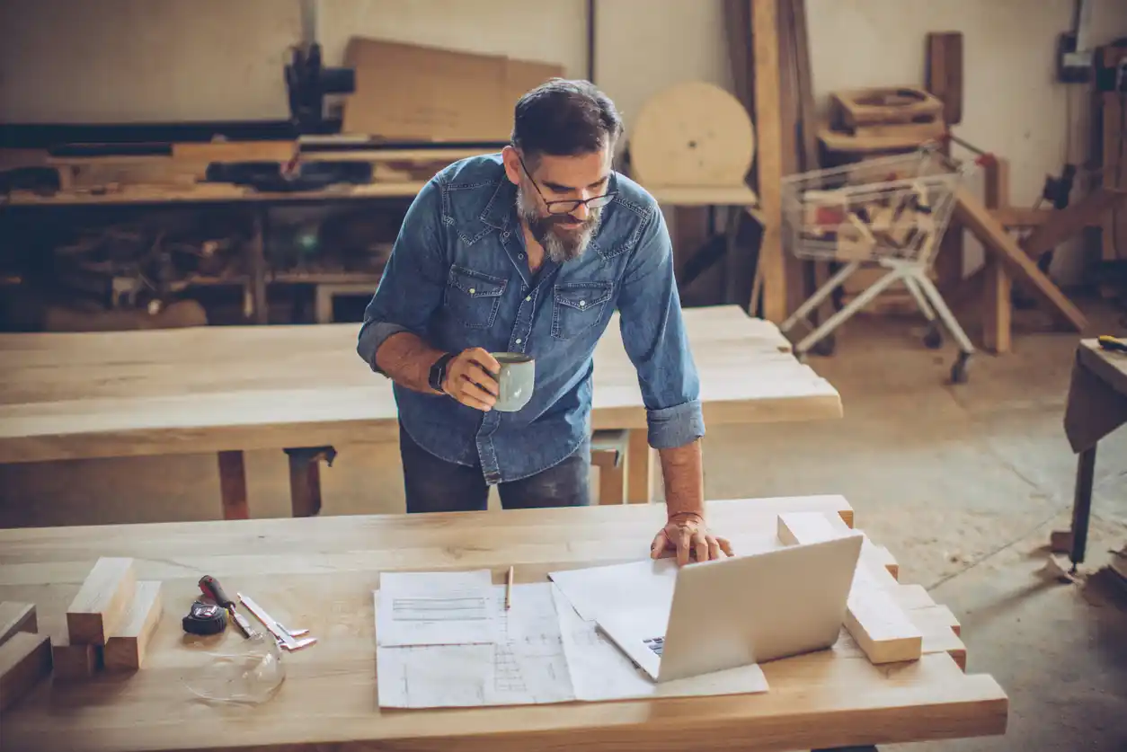 Business owner on laptop with coffee in hand in workshop.