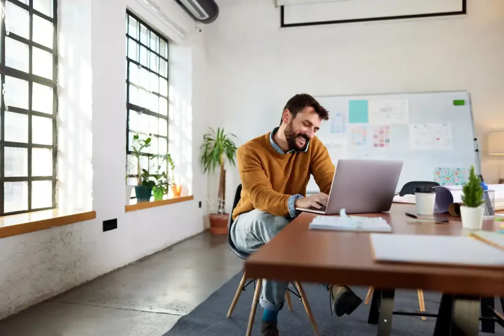 Mortgage broker on laptop smiling in office with email marketing tips on whiteboard behind him.