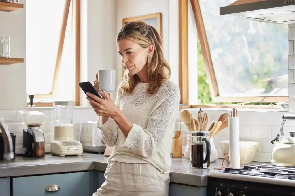 Woman on phone in brightly lit kitchen, holding coffee and phone, searching for mortgage broker online.